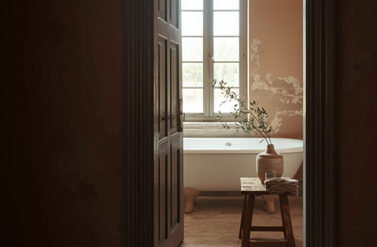 A freestanding bathtub beneath tall windows in a softly lit bathroom, with a wooden stool, folded linens, and an earthenware vase creating a calm, ritual-inspired atmosphere.
