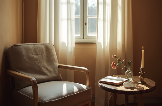A cozy reading corner with a cream armchair beside a sunlit window, a small table holding an open journal, candle, a clear crystal, and fresh roses in a warm, peaceful setting.